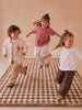 Three young children joyfully run and jump on The Diamond, a Little Forest Animals Silk Rug, in a modern child's room. They wear casual, neutral-toned outfits and have their hair styled in half-up buns. The plain beige background creates a warm, playful atmosphere.