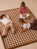 Three young children sit and lie on the Little Forest Animals Silk Rug — The Diamond. One child sits cross-legged, another lies on their back, and the third lays on their stomach, all appearing relaxed and engaged with one another. The scene is set atop this luxury children's rug, which features a subtle diamond harlequin pattern that adds both comfort and elegance to their playful gathering.