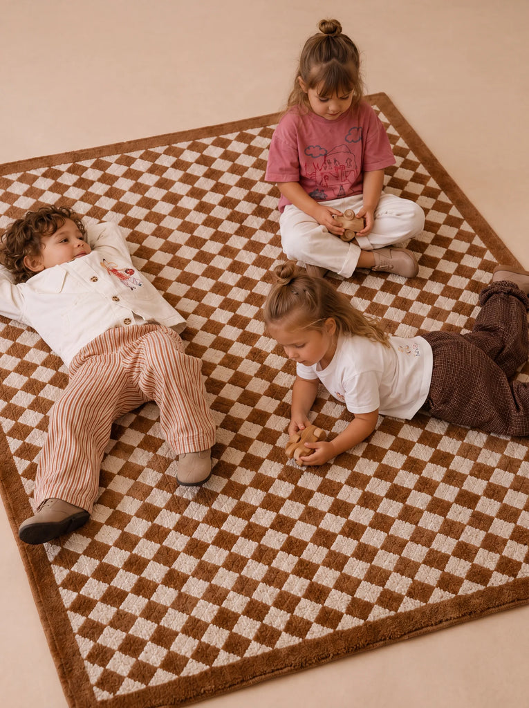 Three young children sit and lie on the Little Forest Animals Silk Rug — The Diamond. One child sits cross-legged, another lies on their back, and the third lays on their stomach, all appearing relaxed and engaged with one another. The scene is set atop this luxury children's rug, which features a subtle diamond harlequin pattern that adds both comfort and elegance to their playful gathering.