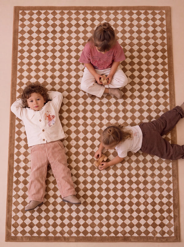 Three children sit and lie on the Little Forest Animals Silk Rug — The Diamond. One child lies on their back with hands behind their head, another sits cross-legged, and a third lies on their stomach playing with a toy atop the rug’s checkered brown and white pattern.