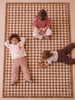 Three children sit and lie on the Little Forest Animals Silk Rug — The Diamond. One child lies on their back with hands behind their head, another sits cross-legged, and a third lies on their stomach playing with a toy atop the rug’s checkered brown and white pattern.
