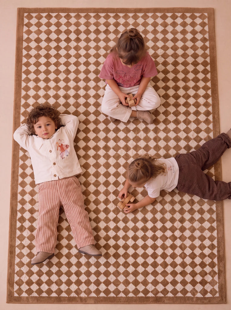 Three children sit and lie on the Little Forest Animals Silk Rug — The Diamond. One child lies on their back with hands behind their head, another sits cross-legged, and a third lies on their stomach playing with a toy atop the rug’s checkered brown and white pattern.