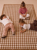 Three young children sit and lie on The Diamond — Little Forest Animals Silk Rug, featuring a beige and white checkered design. Two girls play with toys while a boy relaxes nearby with his hands behind his head, all appearing comfortable and content on this luxurious, hand-tufted children’s rug.