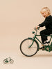A young child in black clothes rides a green bicycle, smiling and looking down at Little Forest Animals — The Sunday Ride (ships mid-September), a die-cast metal model of a vintage city bike, on the floor in front of them against a plain beige background.