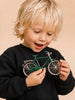 A young child with blonde hair, wearing a black sweatshirt, smiles excitedly while holding and admiring Little Forest Animals — The Sunday Ride, a small handcrafted metal bicycle model in vibrant green.