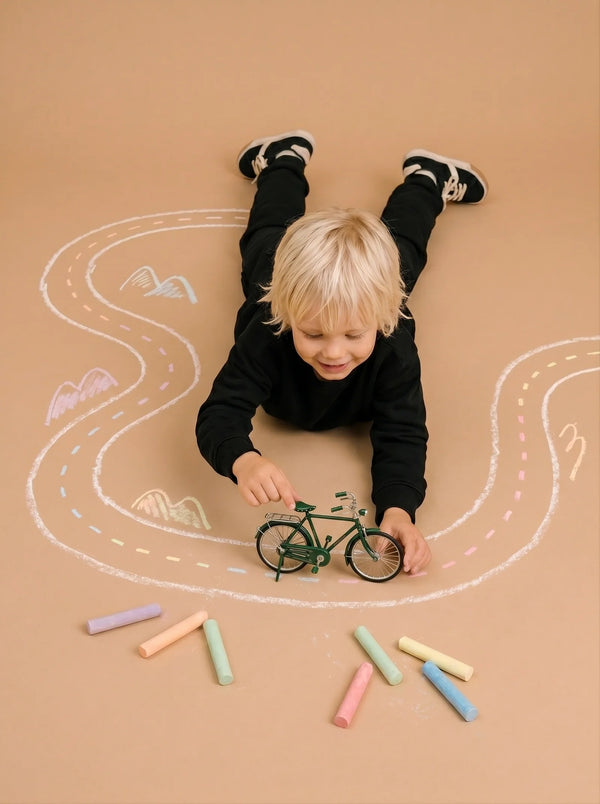 A young child with blonde hair plays with Little Forest Animals — The Sunday Ride, a handcrafted metal bicycle model, on a floor decorated with a chalk-drawn road and colorful pieces of chalk scattered nearby.
