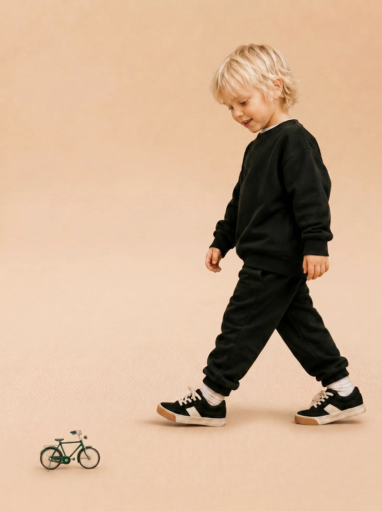 A young child in black sweatpants and a sweatshirt walks on a beige floor, smiling and looking down at Little Forest Animals — The Sunday Ride, a small handcrafted metal bicycle model, in front of them.