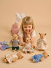A young girl lies on her stomach, smiling at four Little Forest Animals — The Thompson Family dolls dressed in colorful outfits, with extra doll clothes arranged on the floor nearby, all set against a beige background.
