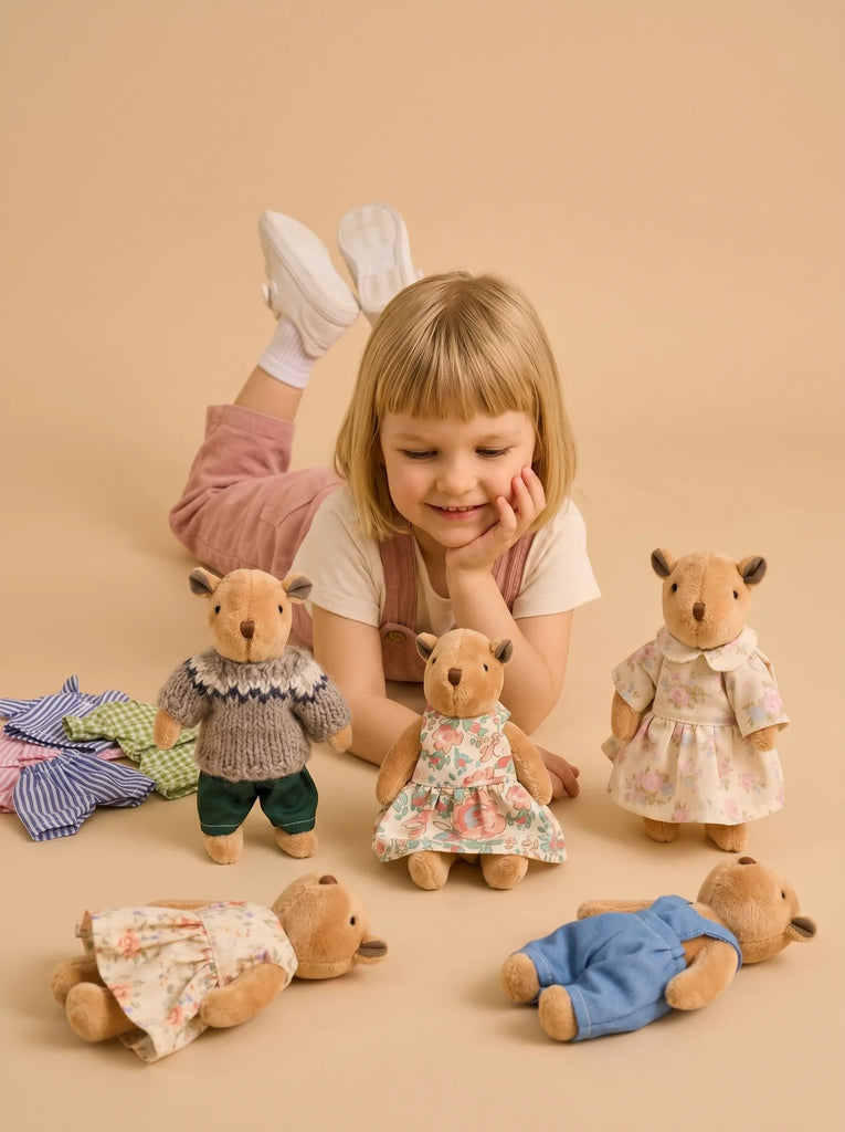 A young girl lies on her stomach, smiling at four Little Forest Animals — The Thompson Family dolls dressed in colorful outfits, with extra doll clothes arranged on the floor nearby, all set against a beige background.