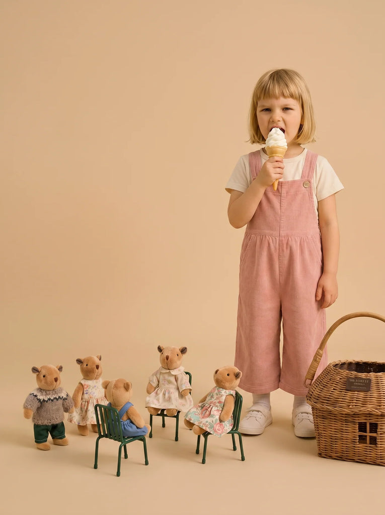 A young girl in pink overalls eats an ice cream cone, standing next to a rattan basket gift set. In front of her are five Little Forest Animals — The Thompson Family figurines, some sitting on small chairs, arranged as if at a tea party.