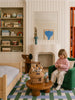 A young child sits on a green chair in a cozy, well-decorated bedroom with a small bed, shelves of toys and books, a fireplace, and a round coffee table holding the Little Forest Animals — The Thompson Family gift set, featuring organic cotton dolls and other cherished stuffed animals.