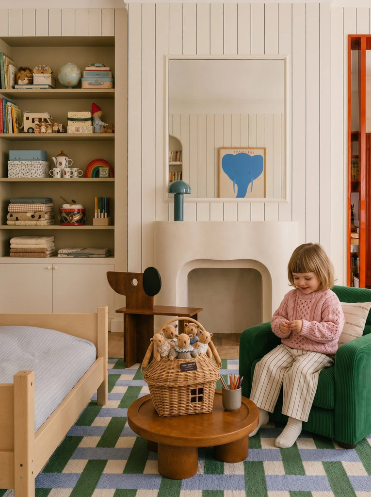 A young child sits on a green chair in a cozy, well-decorated bedroom with a small bed, shelves of toys and books, a fireplace, and a round coffee table holding the Little Forest Animals — The Thompson Family gift set, featuring organic cotton dolls and other cherished stuffed animals.