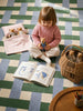 A young child sits on a checkered rug, holding a small plush from the Little Forest Animals — The Thompson Family collection. Nearby, an open picture book rests next to a rattan basket gift set filled with more toys, while two additional Thompson Family stuffed animals are tucked under a blanket.
