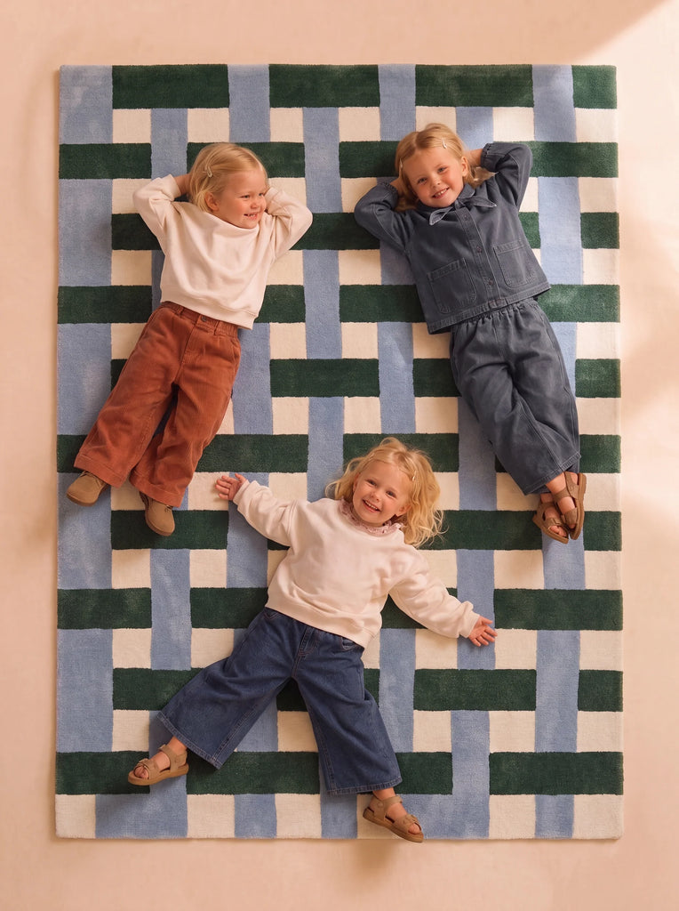 Three young children smile and lie on their backs, arms behind their heads, dressed in casual, comfortable earthy-toned clothing, on The Weave’s Little Forest Animals Silk Rug.