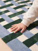 A child's hand with a white sleeve touches the soft surface of The Weave's Little Forest Animals Silk Rug, featuring alternating squares in blue, white, and dark green.