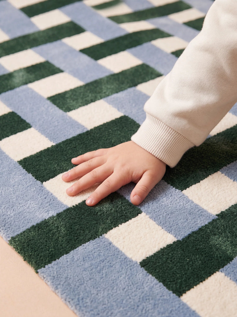 A child's hand with a white sleeve touches the soft surface of The Weave's Little Forest Animals Silk Rug, featuring alternating squares in blue, white, and dark green.