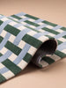 A close-up of the Little Forest Animals Silk Rug — The Weave, featuring a woven pattern in green, blue, and white, partially rolled up on a beige surface.