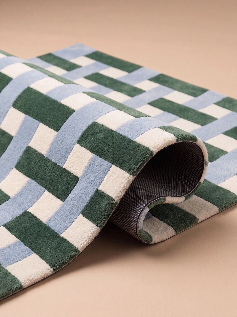 A close-up of the Little Forest Animals Silk Rug — The Weave, featuring a woven pattern in green, blue, and white, partially rolled up on a beige surface.