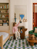A young child sits on a wooden chair in a cozy, colorful bedroom, holding up a teddy bear. Toys, books, and stuffed animals fill the tidy shelves—including the charming Little Forest Animals — The Thompson Family organic cotton dolls—while a rattan basket gift set nestles nearby. A bed and green armchair are arranged on a patterned rug, completing the warm and inviting scene.