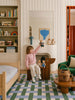 A child sits on a wooden chair in a cozy, colorful bedroom, holding up a teddy bear. Toys, books, and stuffed animals are arranged on shelves and around the room. A basket and bed are also visible atop the Little Forest Animals Silk Rug by The Weave, a beautiful designer area rug that adds warmth and charm to the inviting space.