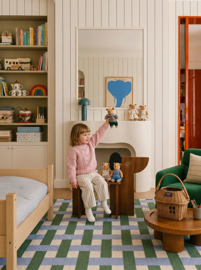 A child sits on a wooden chair in a cozy, colorful bedroom, holding up a teddy bear. Toys, books, and stuffed animals are arranged on shelves and around the room. A basket and bed are also visible atop the Little Forest Animals Silk Rug by The Weave, a beautiful designer area rug that adds warmth and charm to the inviting space.