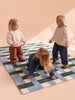 Three young children with blonde hair play together on The Weave’s Little Forest Animals Silk Rug, which features a checkered green, blue, and white designer pattern. Two stand and one crouches, all smiling and interacting in a softly lit room.