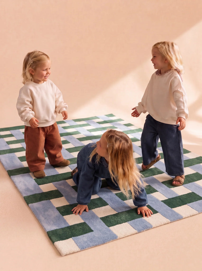 Three young children with blonde hair play together on The Weave’s Little Forest Animals Silk Rug, which features a checkered green, blue, and white designer pattern. Two stand and one crouches, all smiling and interacting in a softly lit room.