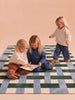Three young children with blonde hair play and sit on The Weave's Little Forest Animals Silk Rug, which features a charming designer pattern. Two of them are reading a book together while the third stands nearby; all are happily engaged in casual clothes.