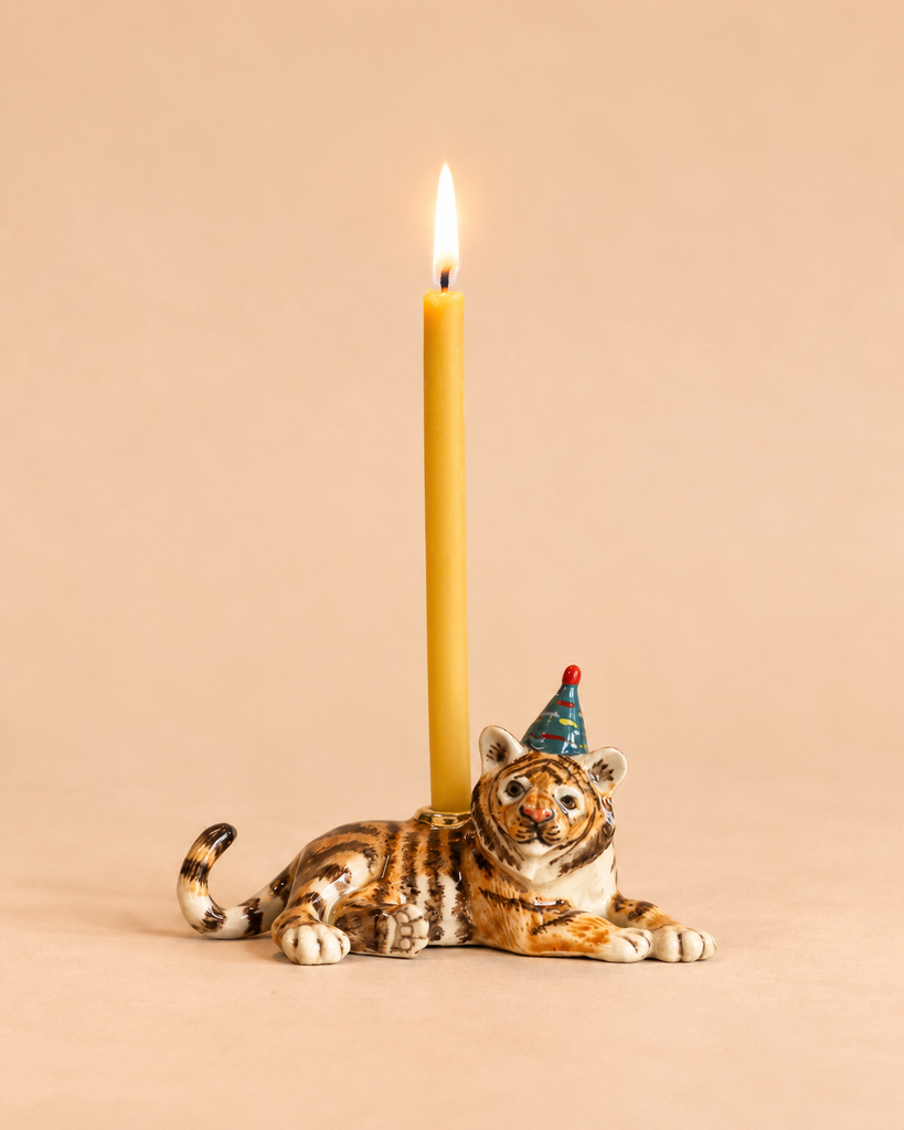 A lit yellow candle is held by the Tiger Cake Topper—a charming, hand-painted ceramic tiger figurine wearing a small blue party hat—set against a plain beige background, making it an adorable birthday decoration.