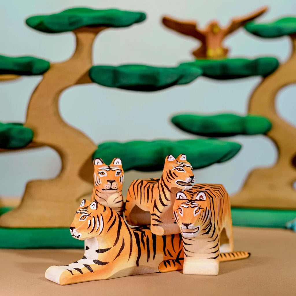 The Bumbu Wooden Tiger Set features handcrafted wooden tiger figurines arranged in a group, with three standing and one lying down. Behind them, crafted wooden trees with green foliage and a wooden owl perched on a branch create a whimsical forest backdrop, perfect for imaginative play.