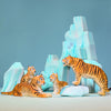 A scene showcases the Bumbu Wooden Tiger Set, consisting of four handcrafted wooden toy tigers—three sitting and one standing—set against a backdrop of icy blue, stylized rocks and mountains. The orange and black-striped tigers seem to be engaging in imaginative play with each other.