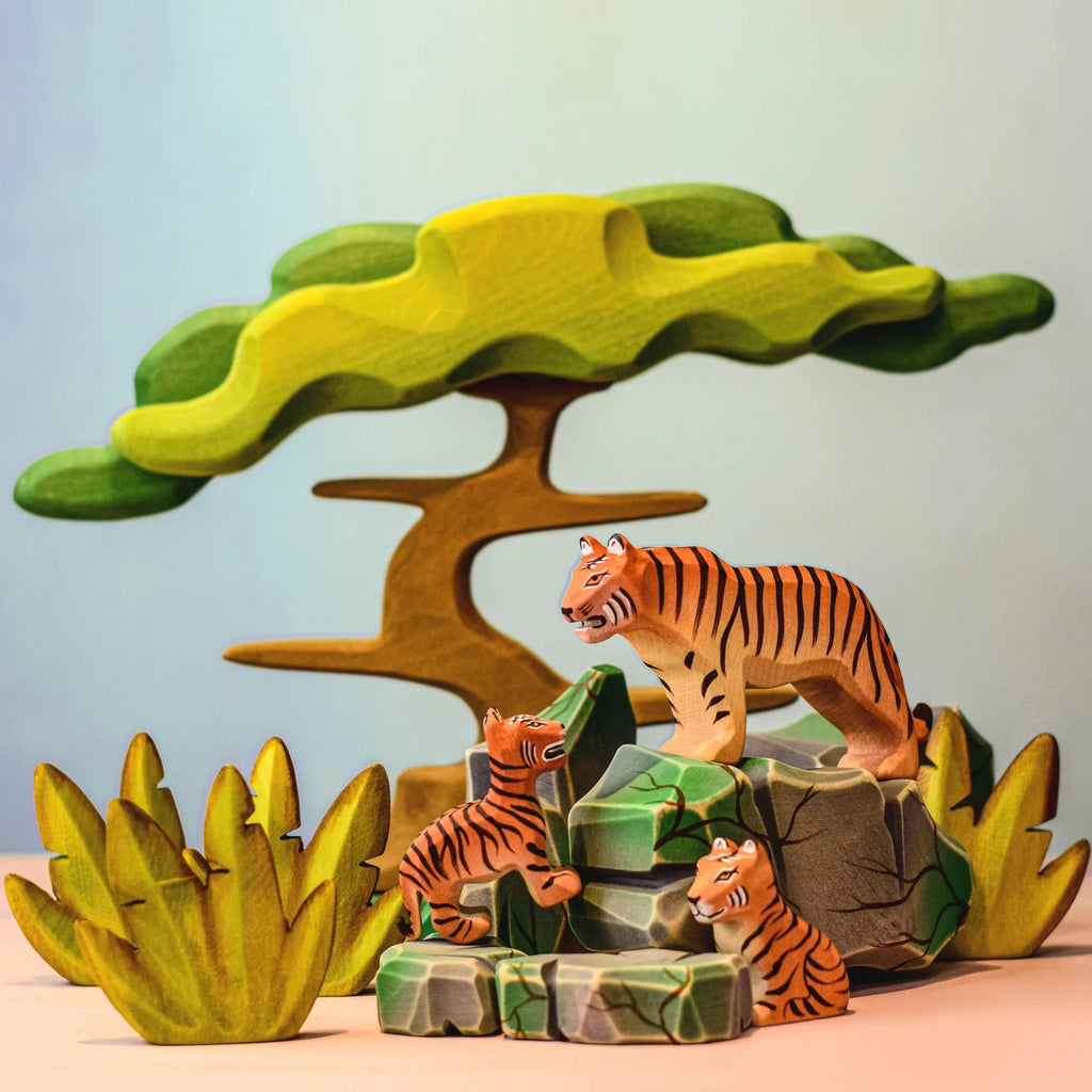 A vibrant depiction showcases the Bumbu Wooden Tiger Set, featuring three meticulously handcrafted wooden tiger figurines in a jungle tableau. Two tiger cubs frolic on rocks, while an adult tiger observes attentively nearby. The backdrop includes a large, stylized green tree and intricately detailed foliage, creating an engaging and lively environment ideal for sparking imaginative play.