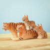 The Bumbu Wooden Tiger Set, displayed on a wooden surface against a light blue backdrop, features four beautifully handcrafted tiger figurines. This set includes one larger reclining tiger, two medium-sized standing tigers, and one smaller sitting tiger, all intricately painted in orange with black stripes—perfect for inspiring imaginative play.