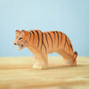 A small wooden figurine of a tiger with an orange body, black stripes, and white details on the face. Handcrafted from eco-friendly materials, this piece is part of the Bumbu Wooden Tiger Set. The figure stands on a light wooden surface against a light blue background.