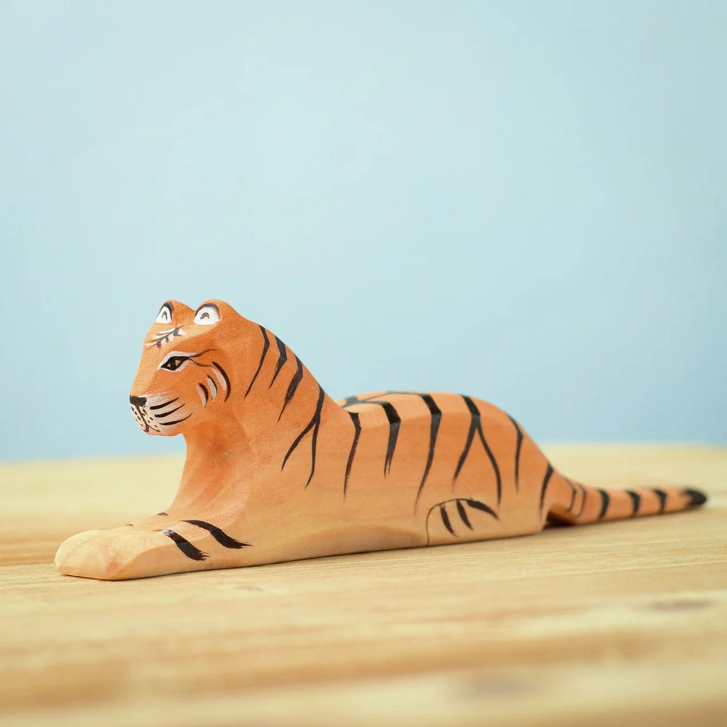 A small, intricately carved wooden tiger figurine from the Bumbu Wooden Tiger Set, featuring painted orange and black stripes. The tiger is lying down on a light wooden surface against a pale blue background. The handcrafted toy showcases detailed eyes, nose, and mouth painted onto eco-friendly wood.