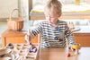 A young boy in a striped shirt uses tongs to sort natural items and purple wooden eggs on the Grapat Tinker Tray, with a wicker basket and Montessori materials encouraging loose-part play in the background.