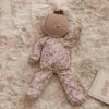 An Olli Ella Dozy Dinkum - Tootsie Briar doll, dressed in a purple floral onesie, rests on a floral-patterned blanket beside a small bouquet of white flowers and a card featuring a bunny illustration.