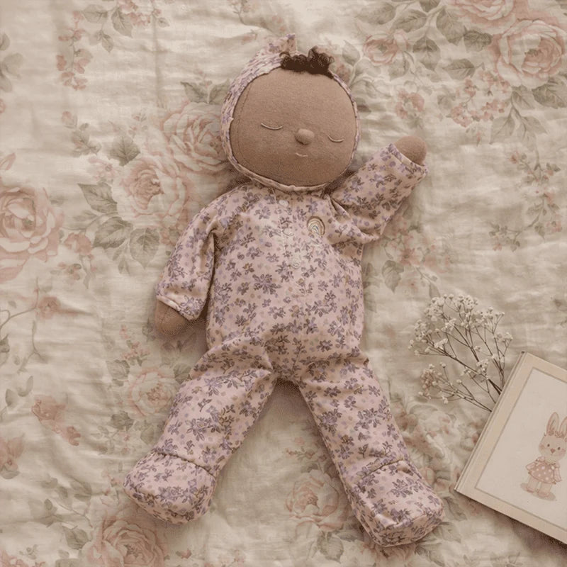 An Olli Ella Dozy Dinkum - Tootsie Briar doll, dressed in a purple floral onesie, rests on a floral-patterned blanket beside a small bouquet of white flowers and a card featuring a bunny illustration.