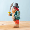 The Bumbu Handmade Wooden Treasure Hunt Set features a handcrafted pirate figurine with a skull hat, eyepatch, red pants, green coat, and yellow-handled sword posed on wood against a light blue background.