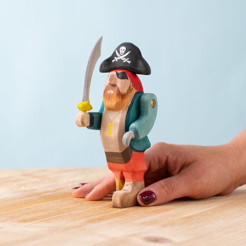 A hand holds a pirate figure from the Bumbu Handmade Wooden Treasure Hunt Set, featuring a sword, eyepatch, and black hat with skull and crossbones, displayed on a light wooden surface with a pale blue background.