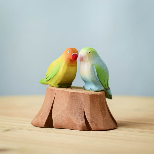 Two vibrant wooden birds from the Bumbu Handmade Wooden Tree Stump and Lovebirds Set rest side by side on a carved tree stump, set against a light blue background—perfect handcrafted nature toys for imaginative play.