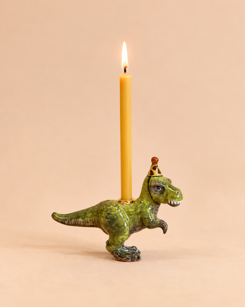 A yellow candle is lit and held by the “T-Rex Cake Topper - Final Sale,” a hand-painted ceramic dinosaur figurine wearing a small crown, set against a plain beige background—a whimsical collectible keepsake.