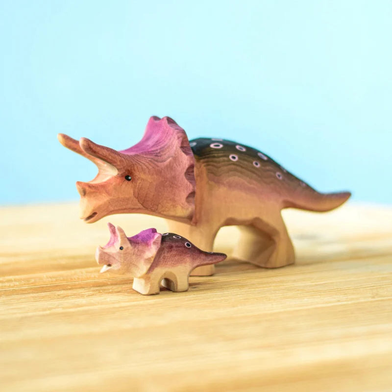 Two wooden toy dinosaurs, resembling triceratops, are displayed on a light wooden surface with a blue background. Handmade from sustainably sourced wood, one is larger with pink accents on the head and back, and the other is smaller with similar pink and purple detailing. Both are painted with non-toxic paint. This is the Bumbu Handmade Triceratops Dinosaur Set.