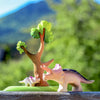 A Bumbu Handmade Triceratops Dinosaur Set depicts an adult and baby near a tree. The background is blurred greenery, suggesting a natural environment. The figures, painted in soft colors with non-toxic paint and fine details, are handmade wooden toys set on a flat green base.