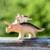 A Bumbu Handmade Triceratops Dinosaur Set features a wooden figurine of a triceratops carrying a smaller triceratops on its back. Detailed with light and dark non-toxic paint, these charming toys rest on a wooden surface, complemented by a blurred green background.