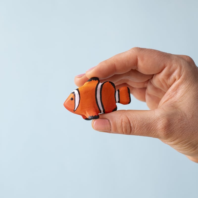 A hand holds a small orange clownfish toy from the Bumbu Handmade Wooden Tropical Fish Set against a plain light blue background.