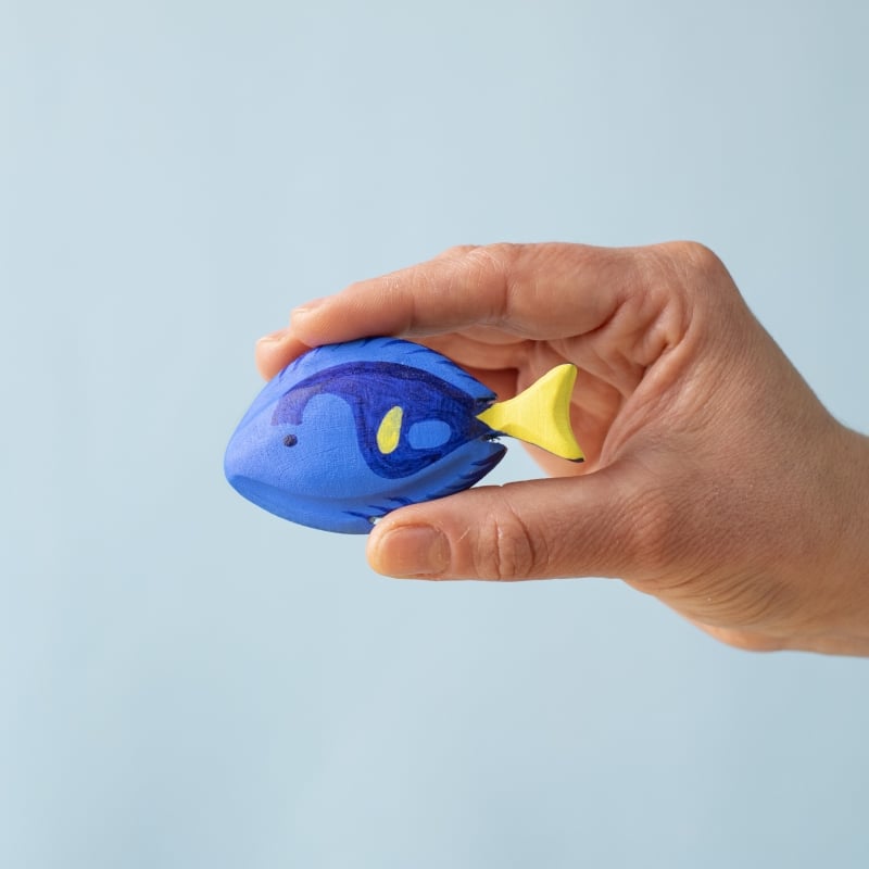 A hand holds a small blue and yellow fish from the Bumbu Handmade Wooden Tropical Fish Set, shown against a light blue background.