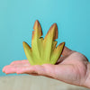A hand holds a Wooden Banana Bush flower, painted with non-toxic paint, highlighting its vibrant green and brown tips against a soft blue background.