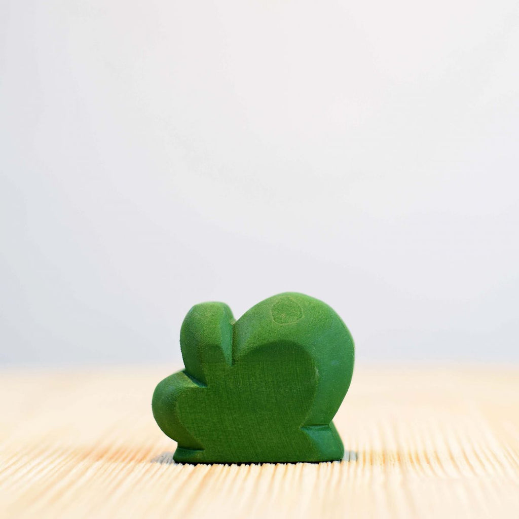 A small green bush-shaped figurine made of wood, painted with non-toxic paint, stands on a light wooden surface with a soft-focus white background.