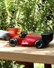 A Playforever Car TURBO Verve Laser Rich Red toy with "turbo" on the side and "SUNRAY" tires sits outdoors on wood, as another blurred racing car toy zooms by in the background amid green foliage.